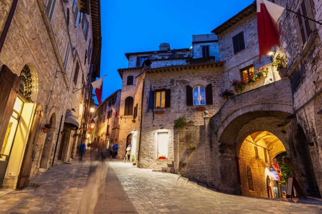 Un vicolo di Assisi all'ora blu con i pennoni bianco-rossi, i lampioni accesi e i gerani alle finestre