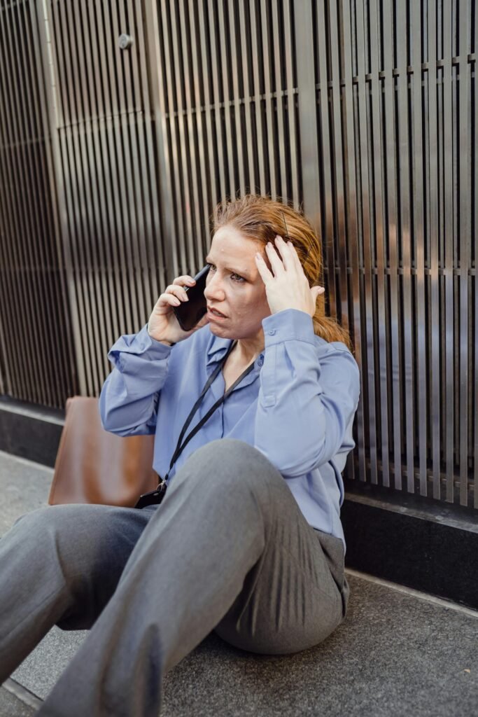 Una donna seduta sul marciapiede in una strada di città, telefono in mano, con espressione preoccupata